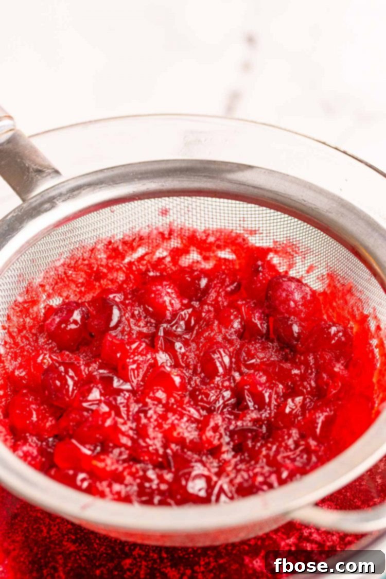 Ruby Red Elixir 5 Cranberry simple syrup mixture being poured through a fine mesh strainer into a large bowl, separating the liquid from the pulp.