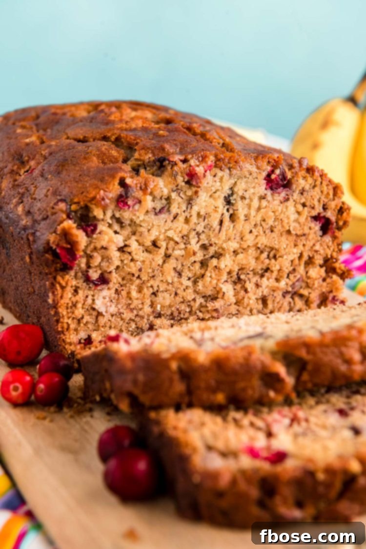 Close up photo of a loaf of cranberry banana bread that had been sliced halfway and is resting on a wooden cutting board.