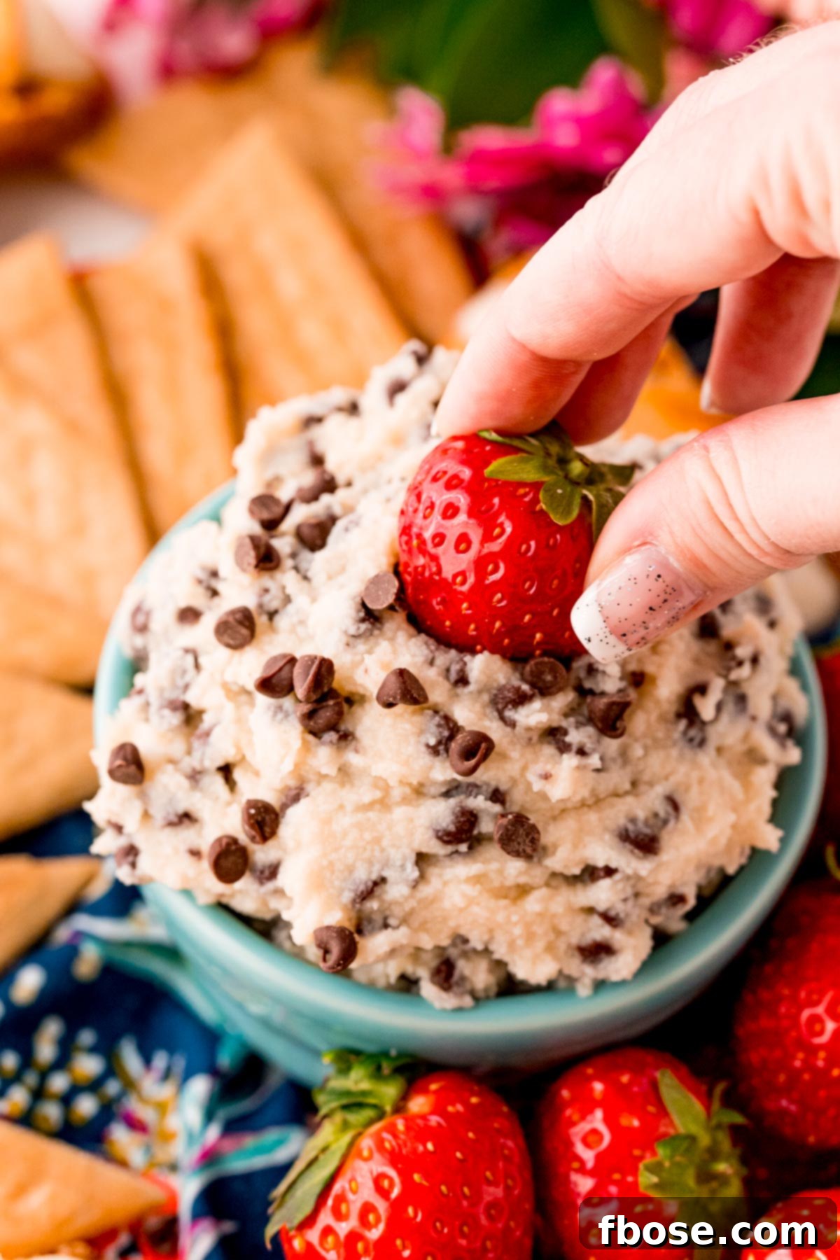 A woman's hand gracefully dipping a fresh strawberry into a luscious dessert dip generously sprinkled with mini chocolate chips, set in a teal bowl.