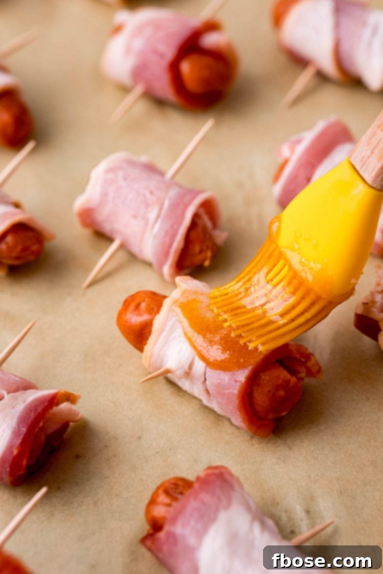 Close up photo of bacon wrapped little smokies on parchment paper being brushed with sauce.