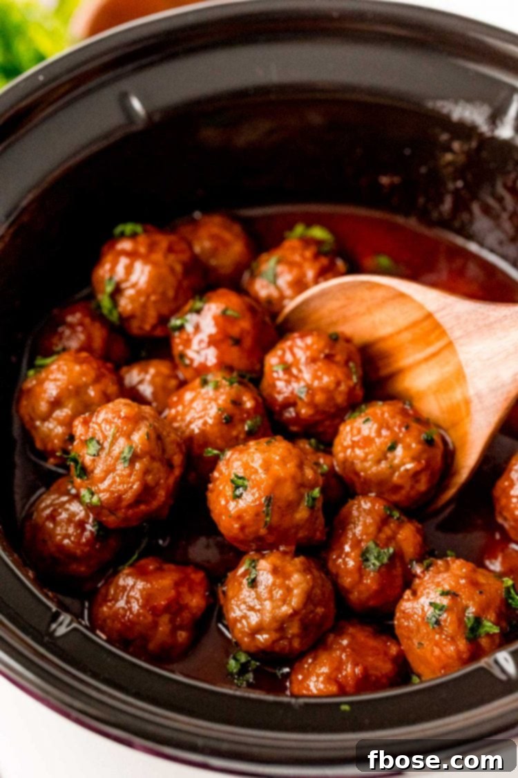 A wooden spoon stirring grape jelly meatballs in a crockpot.
