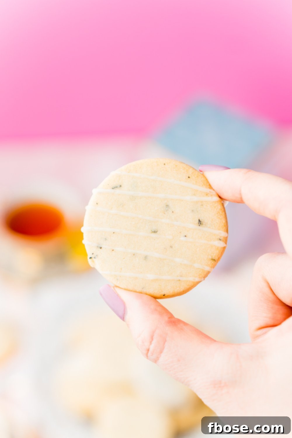 A woman's hand gracefully holds a single Earl Grey Shortbread Cookie, showcasing its delicate round shape and golden hue, with a subtle hint of its lemon glaze.