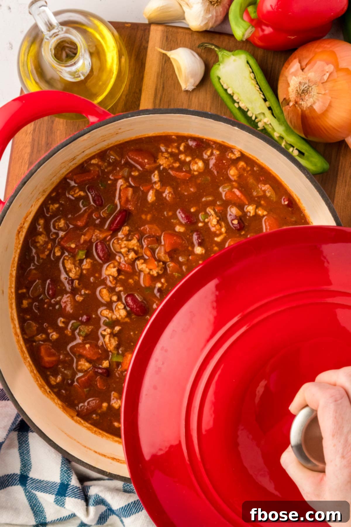 A woman's hand removing the lid of a red Dutch oven to reveal the perfectly cooked turkey chili inside.