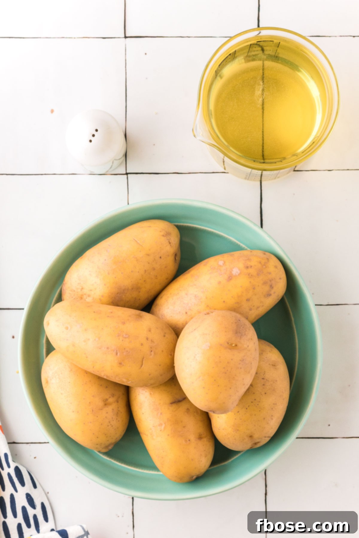 Overhead photo of fresh potatoes, a bottle of vegetable oil, and a small bowl of salt, which are the main ingredients for homemade French fries.