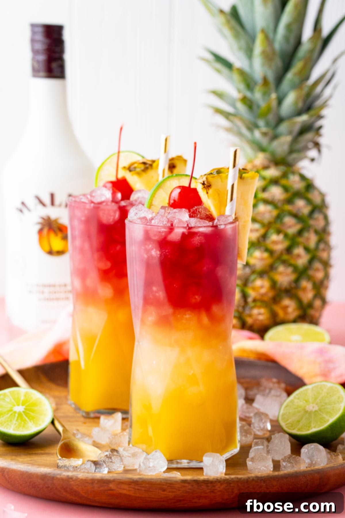Straight on photo of two Malibu Bay Breeze cocktails on a wooden tray with a pineapple and bottle of Malibu rum in the background, showcasing an inviting tropical scene.