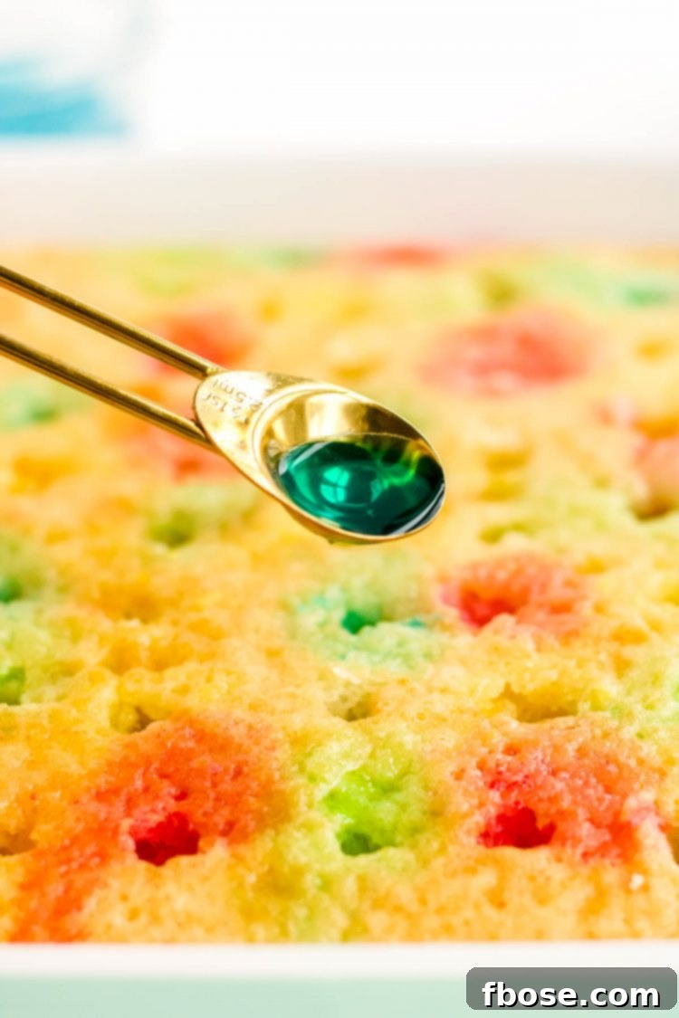 Close up photo of a teaspoon carefully pouring colorful liquid jello into holes on a white poke cake.