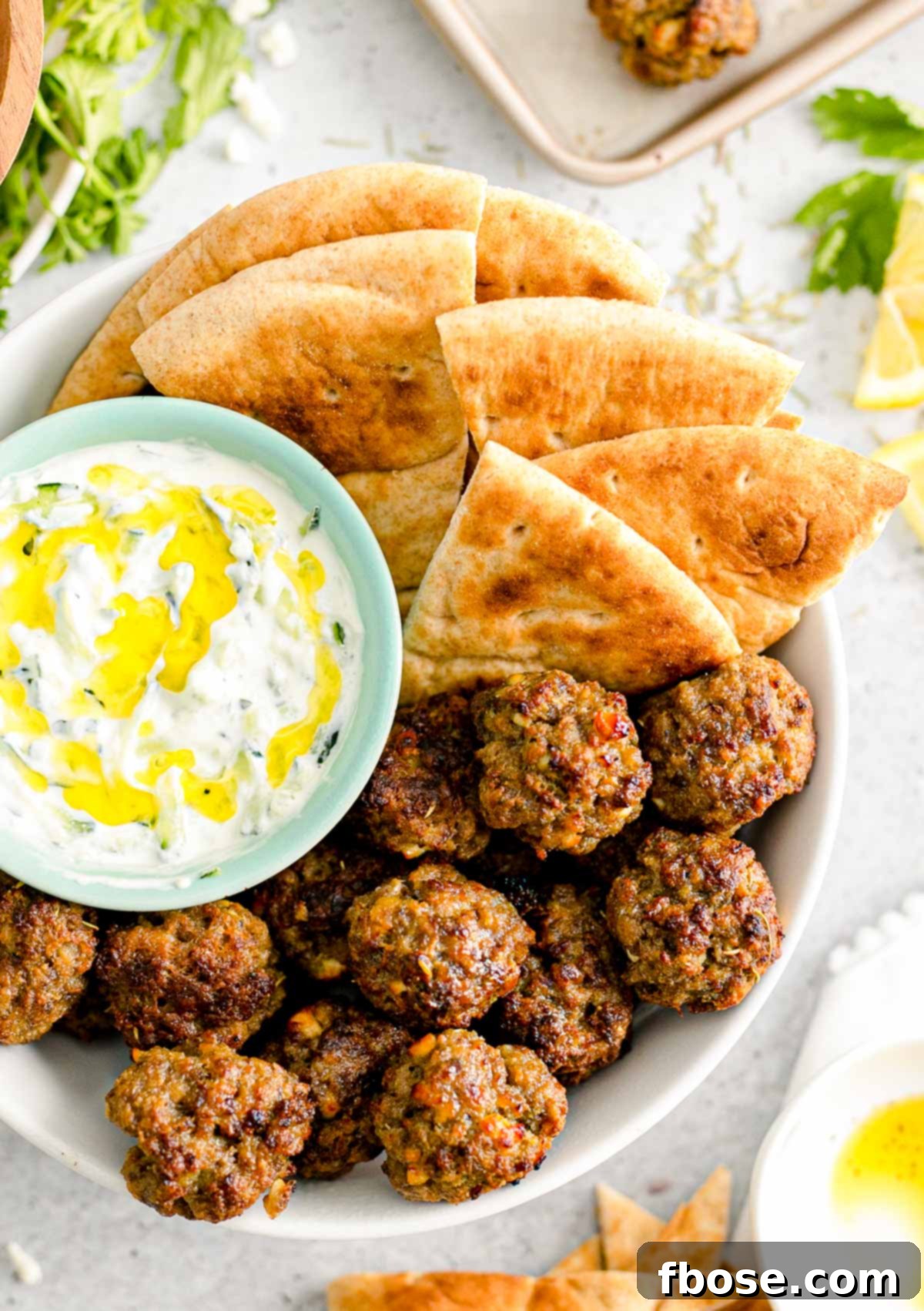 Overhead close up photo of Greek meatballs in a bowl with pita bread and tzatziki sauce, garnished with fresh herbs.