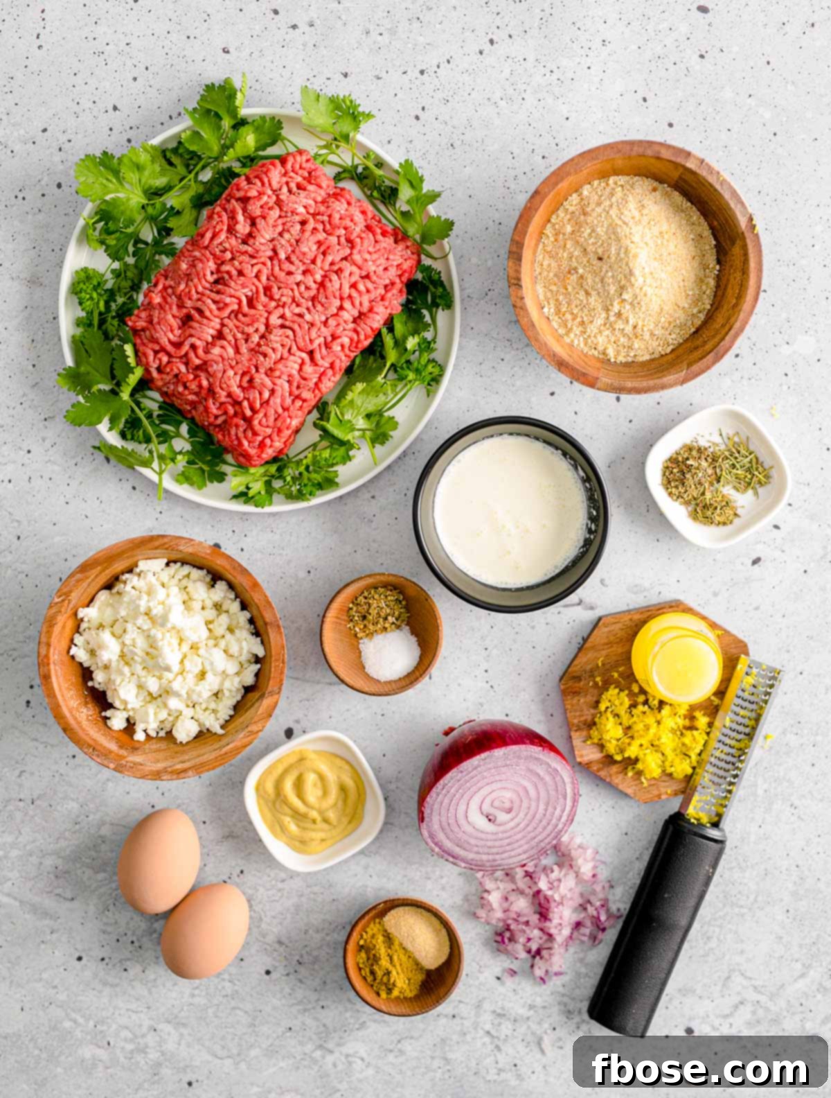 Ingredients to make Greek meatballs arranged neatly on a gray counter top, including ground meat, herbs, cheese, and lemon.