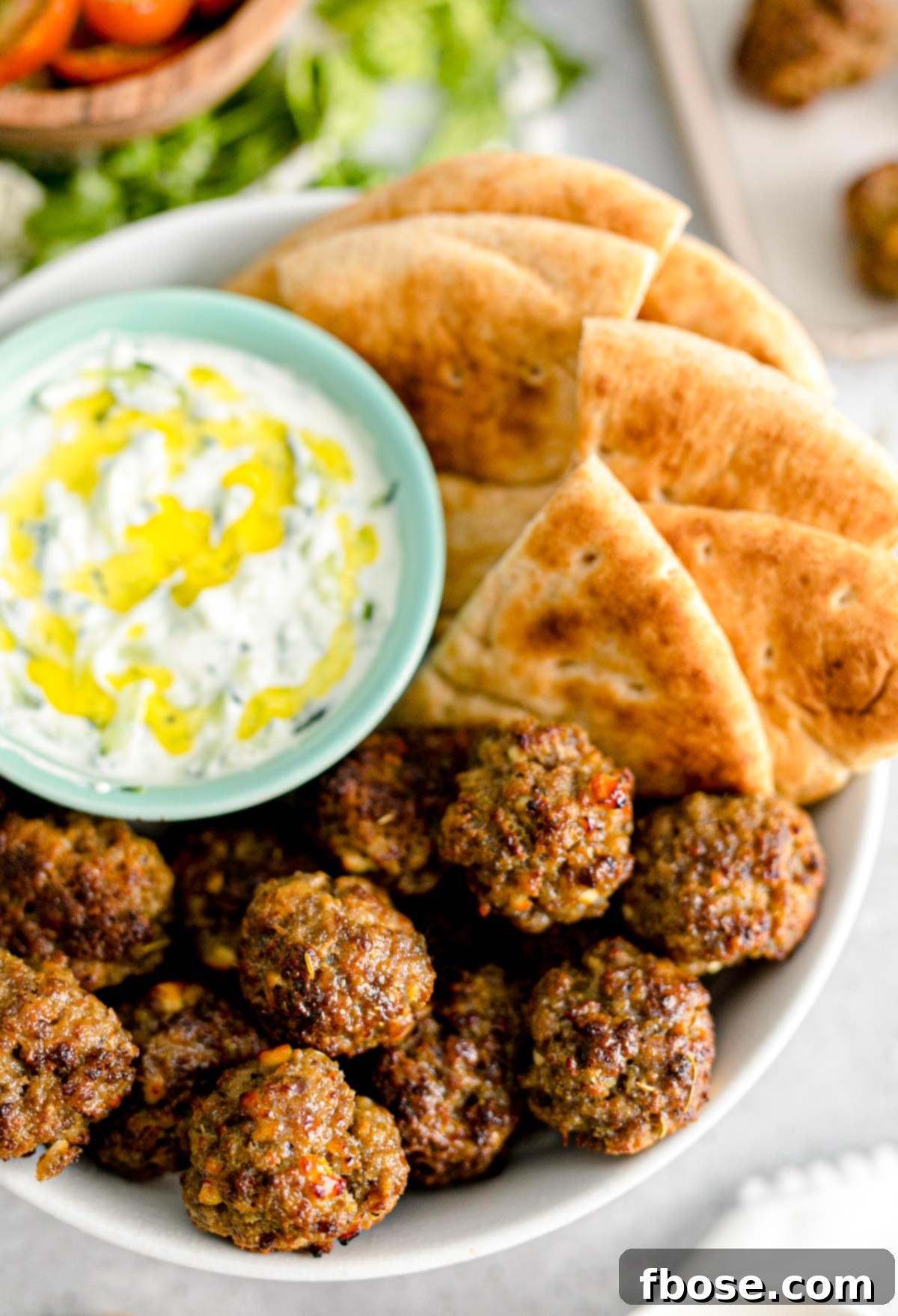 Close up photo of Greek meatballs in a white bowl with tzatziki sauce and a piece of pita bread, garnished with fresh herbs.
