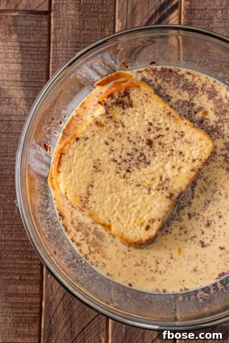 Overhead photo of a slice of brioche bread being dipped in a rich, creamy egg wash, ready for cooking.