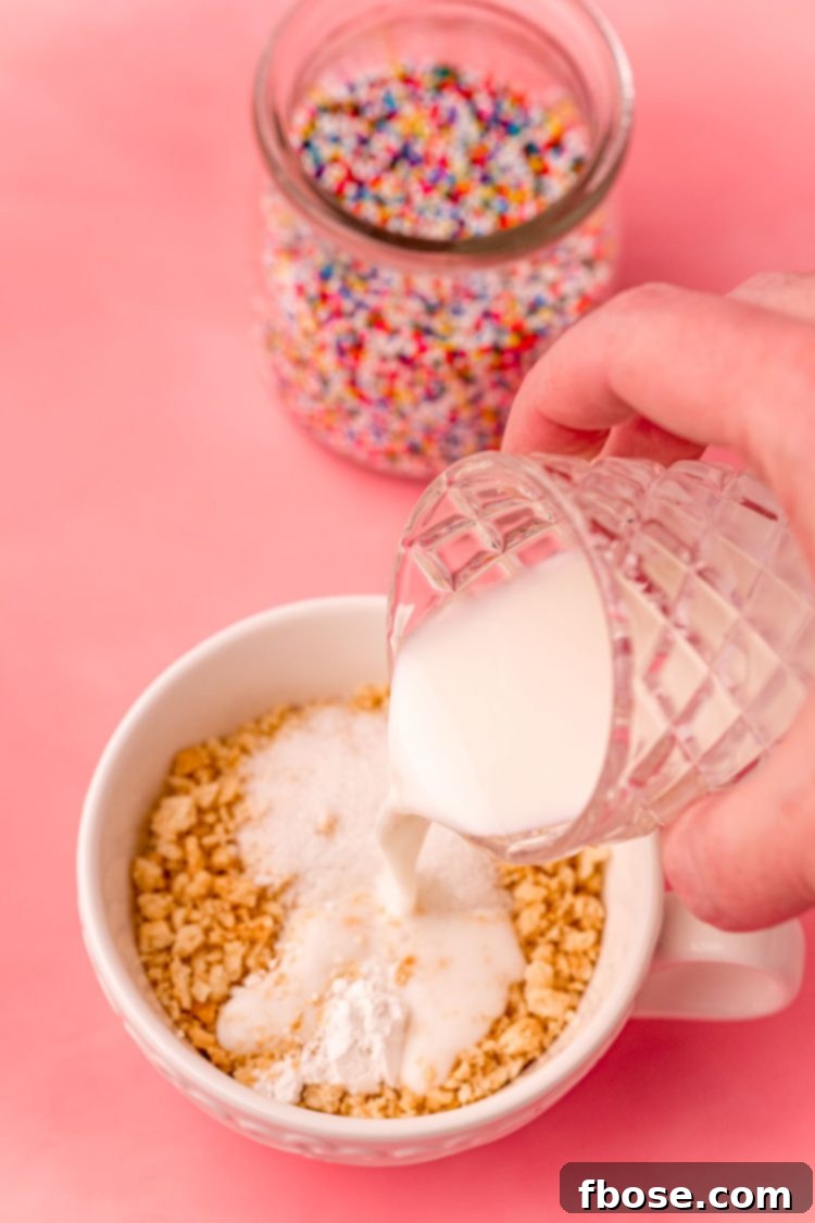 Golden Oreo Confetti Mug Cake 5 A woman's hand pouring milk into a white tea cup with crushed oreos on a pink surface.