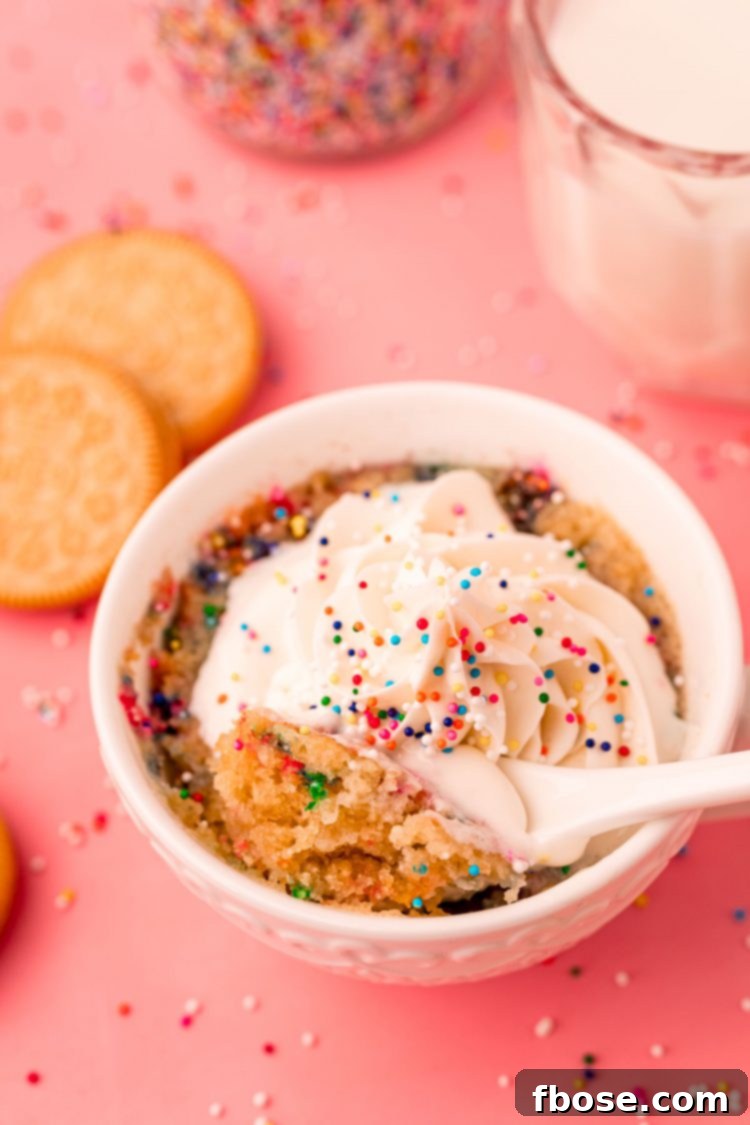 Golden Oreo Confetti Mug Cake 6 A spoon taking a bite out of a golden Oreo mug cake in a white cup on a pink surface.