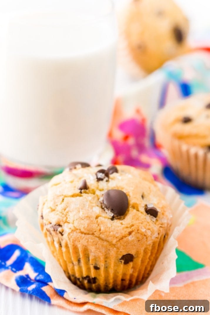 Close up photo of a chocolate chip muffins with a glass of milk and more muffins in the background.