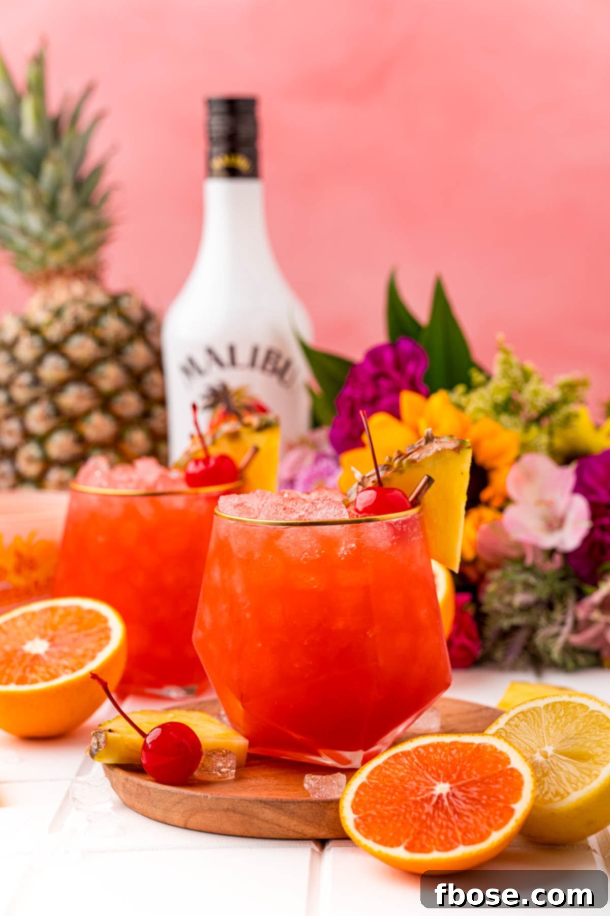Two Bahama Mama cocktails on a wooden board on a white table with a bottle of rum in the background and oranges and pineapple around.