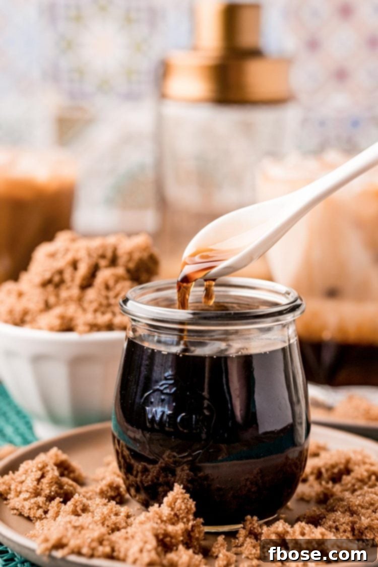 A close-up of a spoon drizzling rich brown sugar simple syrup from a clear glass jar.