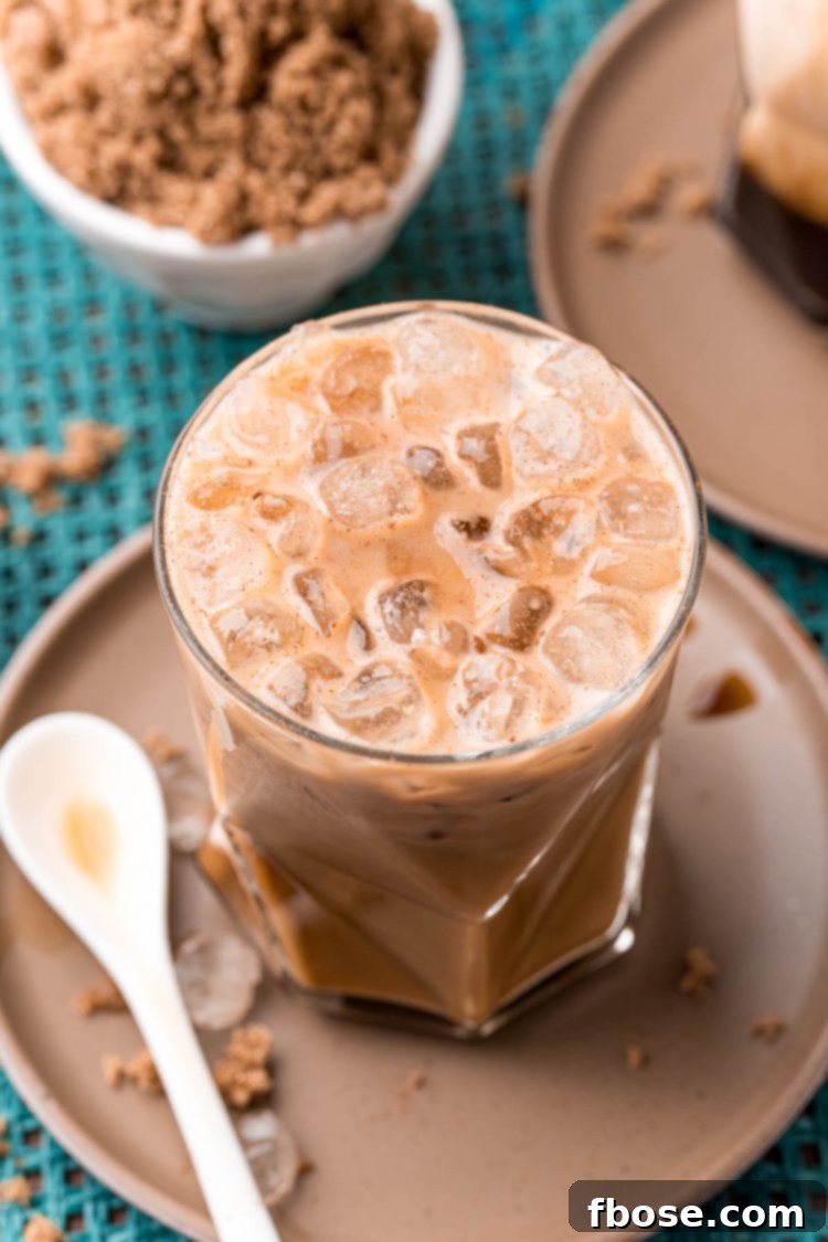 Close up photo of a brown sugar oatmilk shaken espresso on a brown plate with cinnamon sticks and brown sugar. The drink is in a tall glass, garnished with cinnamon, beside whole cinnamon sticks and loose brown sugar.