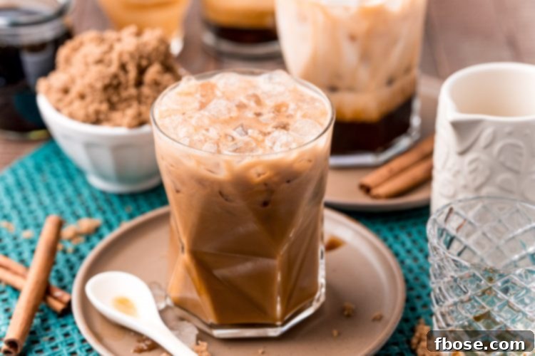 Close up photo of a brown sugar oatmilk shaken espresso on a brown plate with cinnamon sticks and brown sugar. The drink is in a shorter, wider glass with a straw, beside loose brown sugar.