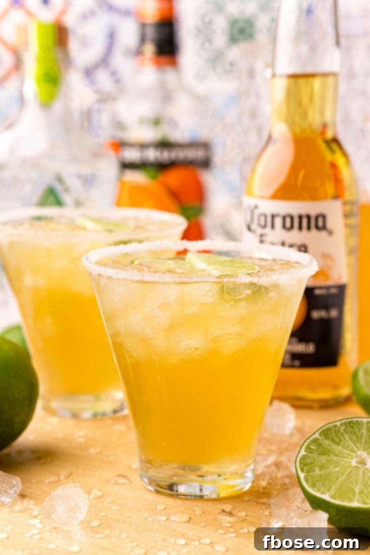 Close-up photo of two Beer Margaritas on a rustic wooden cutting board, surrounded by fresh limes and blurred beer bottles in the background, hinting at the ingredients.