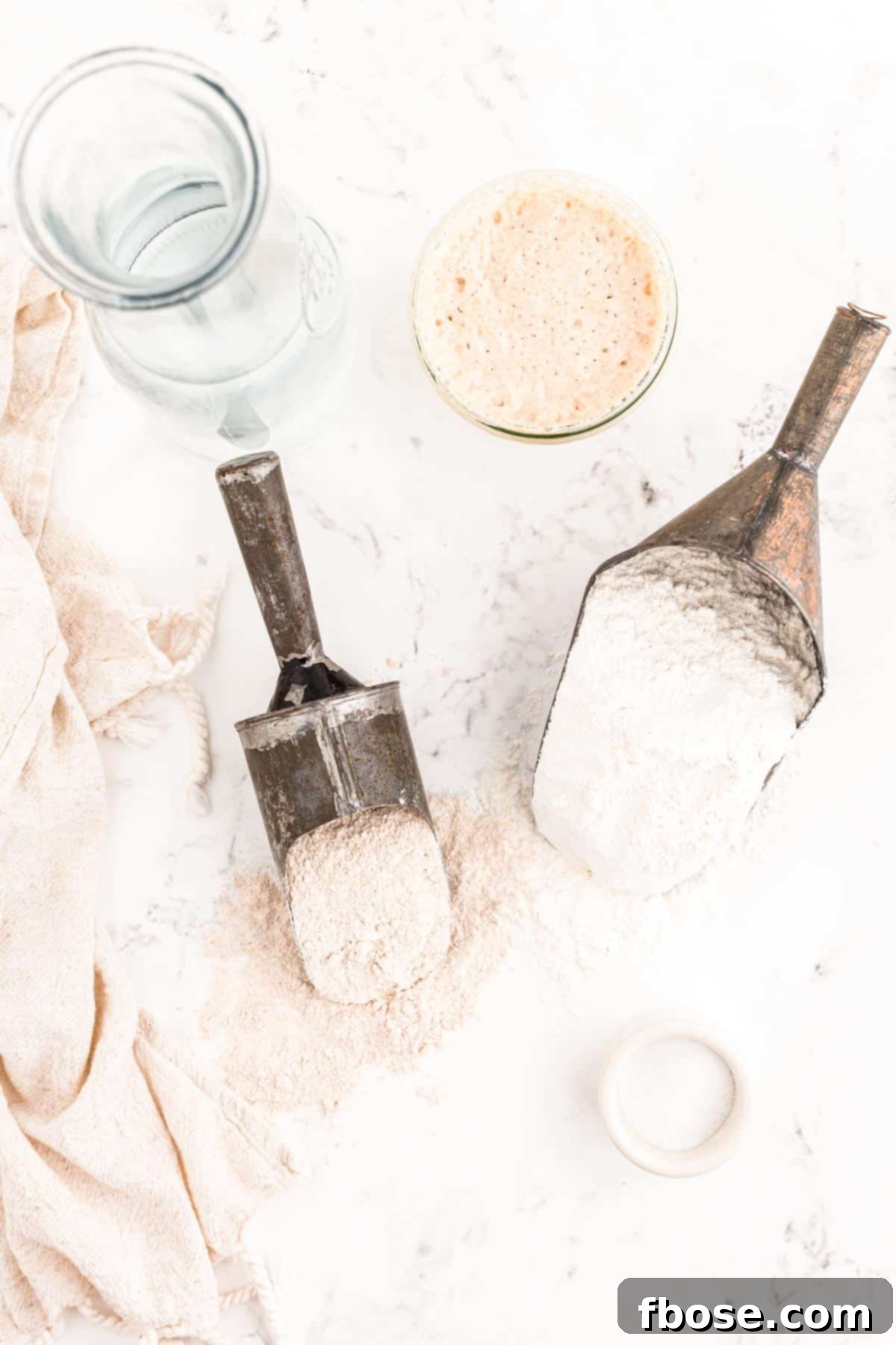 Ingredients for making sourdough bread laid out on a marble countertop, including various flours, water, salt, and starter.