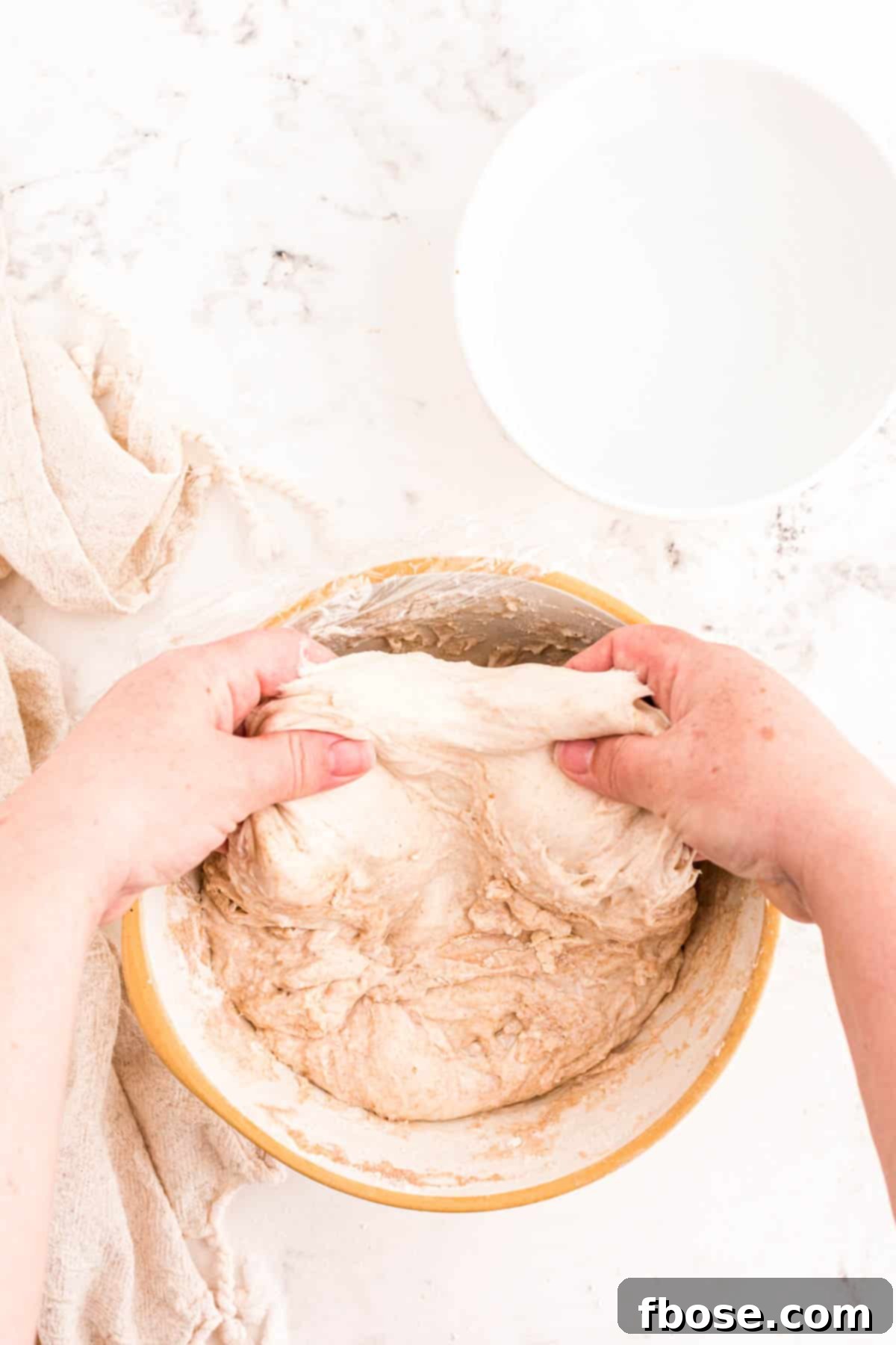 A baker's hand gently reaching under the side of sourdough dough in a bowl to begin a stretch and fold.