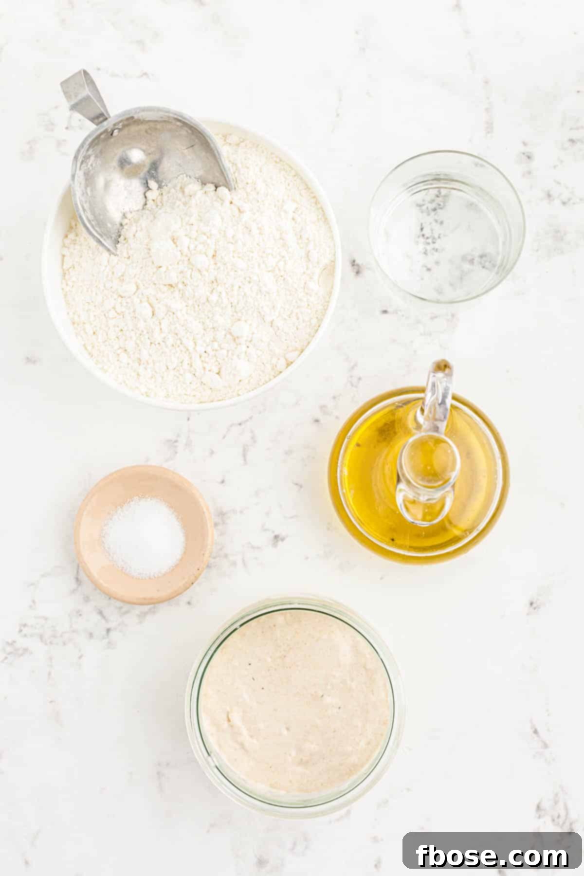 Perfect Sourdough Pizza Dough 4 Various ingredients for making sourdough pizza crust laid out neatly on a pristine marble counter, including flour, water, olive oil, salt, and a jar of sourdough starter.