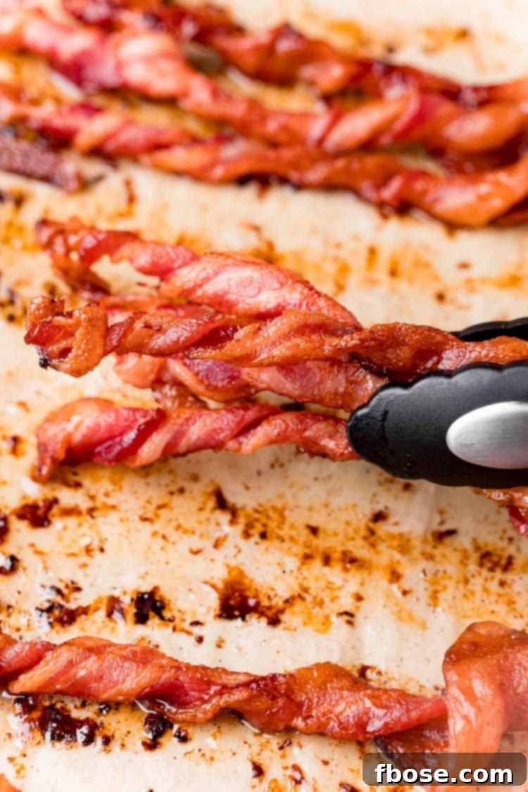 Close up photo of a pair of tongs grabbing twisted bacon off a baking sheet.