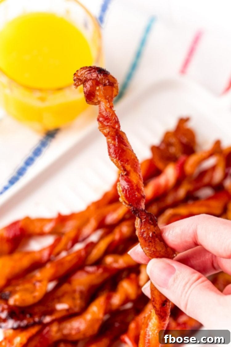 A woman's hand holding a piece of twisted bacon with more bacon and a glass of orange juice in the background.