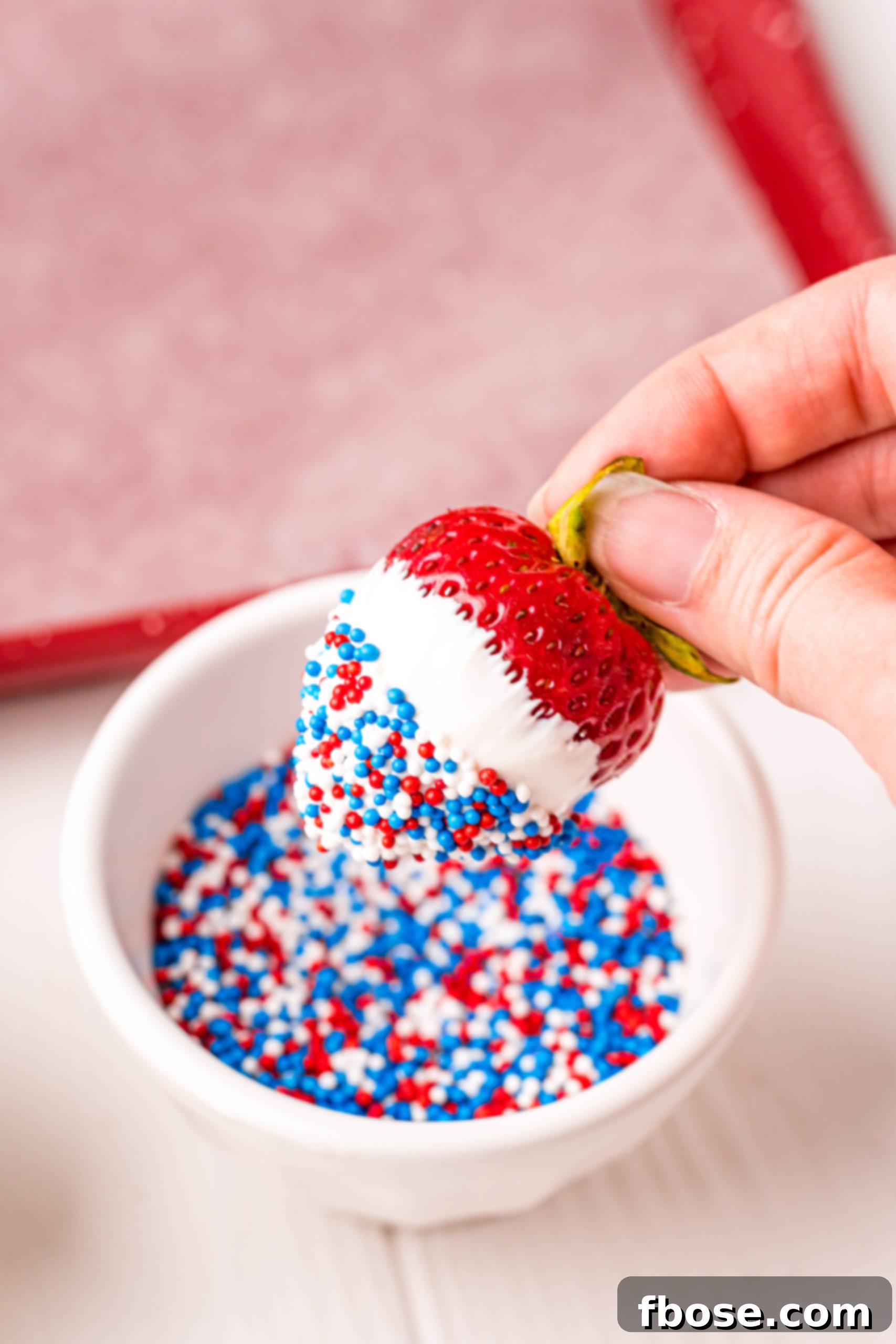 Independence Day Strawberry Delights 5 A woman's hand dipping a white chocolate covered strawberry in red, white, and blue sprinkles.