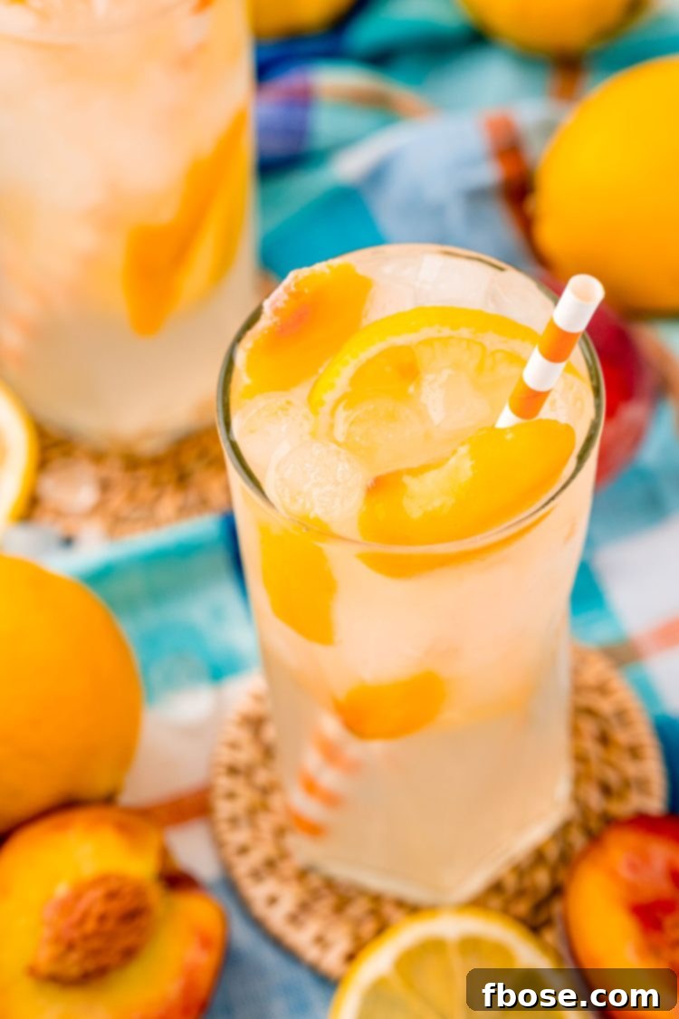 Close up photo of a glass with peach lemonade with lemons and peaches in it and an orange striped straw.