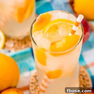 Close up photo of a glass with peach lemonade with lemons and peaches in it and an orange striped straw.