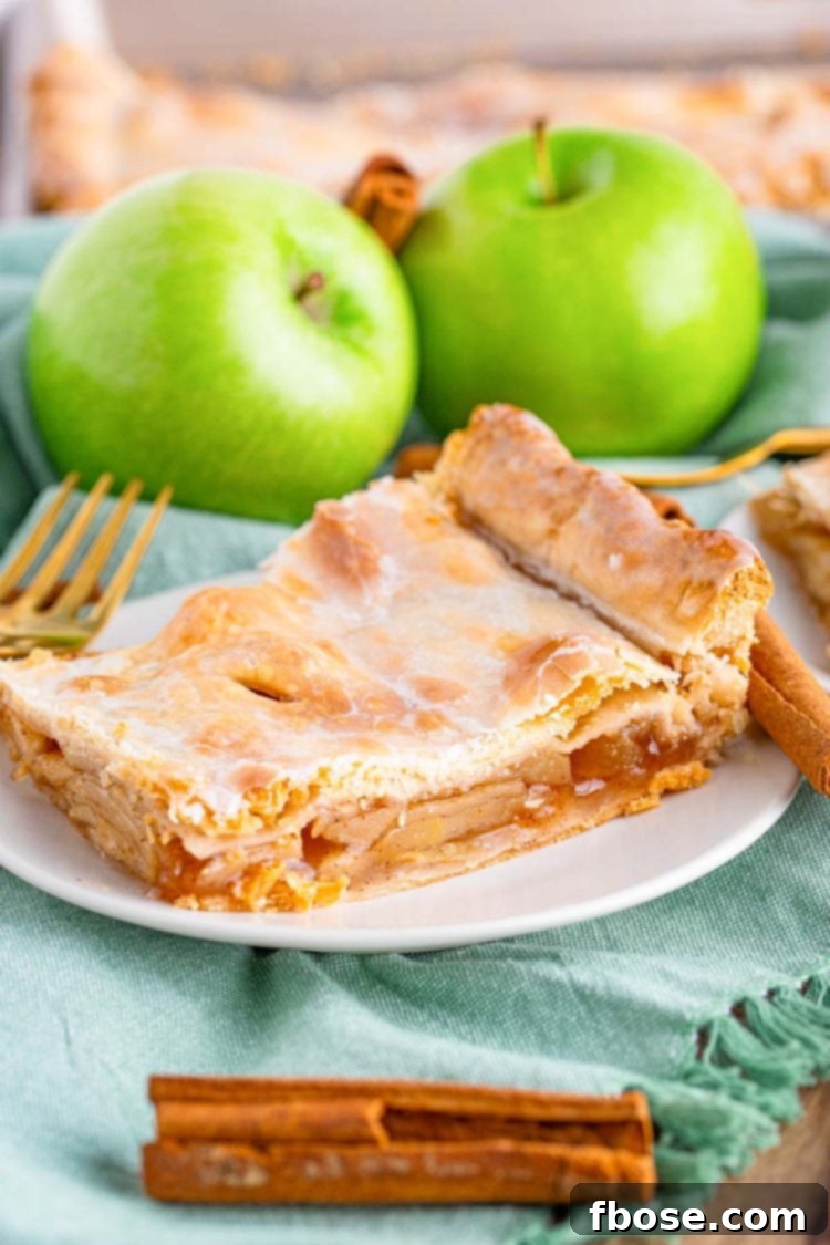 Close up photo of a piece of apple slab pie on a small white plate on a sage green napkin with cinnamon sticks and granny smith apples in the background.