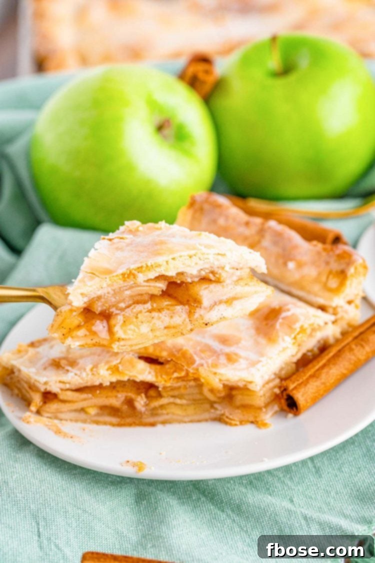 Close up photo of a slice of apple slab pie on a white plate on a blue napkin with a gold fork taking a bite from it. 