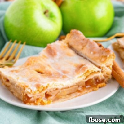 Close up photo of a piece of apple slab pie on a small white plate on a sage green napkin with cinnamon sticks and granny smith apples in the background.