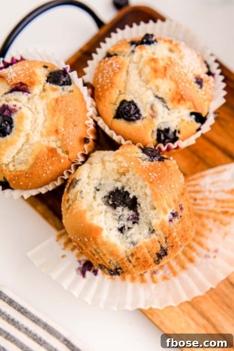 Bakery-Style Jumbo Blueberry Muffins 3 A close-up of three jumbo blueberry muffins on a wooden tray, one with a bite taken out, revealing its moist interior and abundant blueberries.