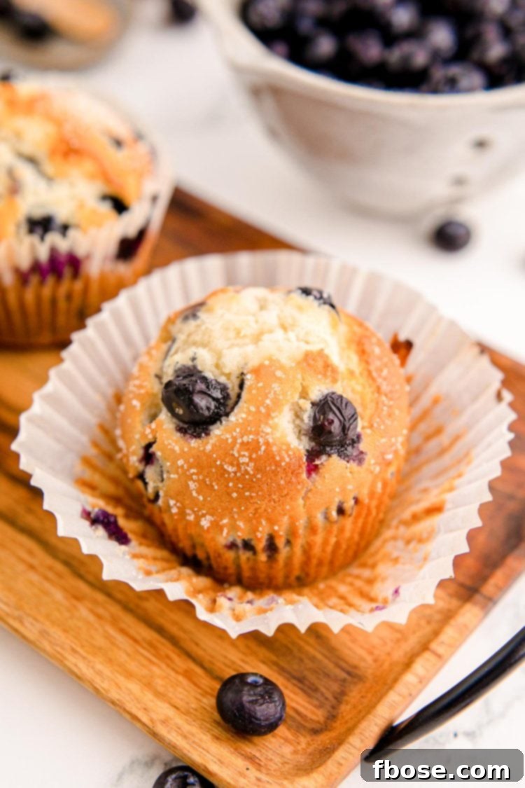 Bakery-Style Jumbo Blueberry Muffins 6 A single jumbo blueberry muffin on a wooden tray with its paper liner partially peeled away, revealing the soft muffin inside.