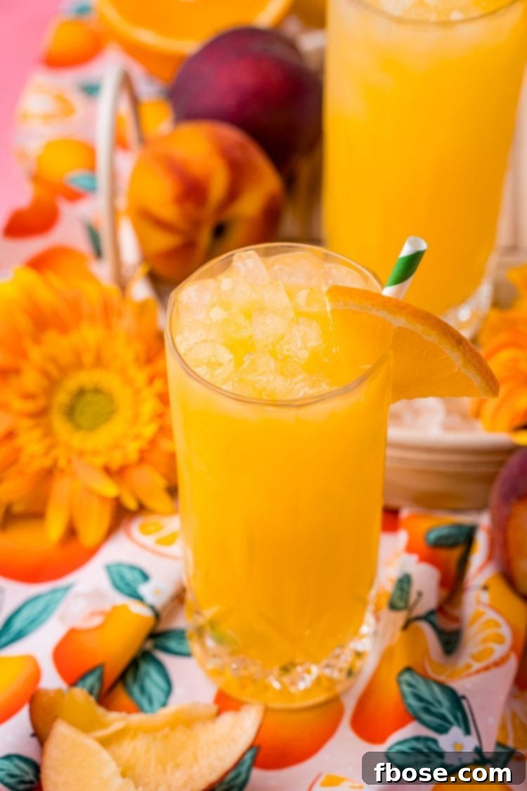 Close up photo of a fuzzy navel drink in a tall glass, garnished with fresh peach and orange slices, on a rattan tray.