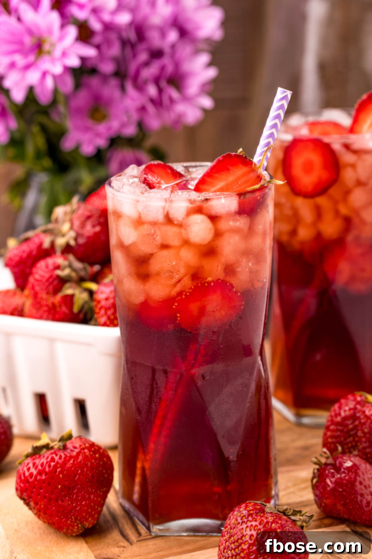 Close up photo of a glass of strawberry tea with another glass and a carton of strawberries in the background, highlighting the freshness of the ingredients.