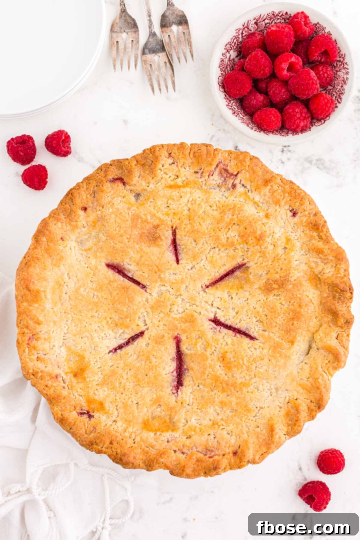 Overhead photo of a whole raspberry pie on a white table with fresh raspberries scattered around it, showcasing its perfect golden crust.