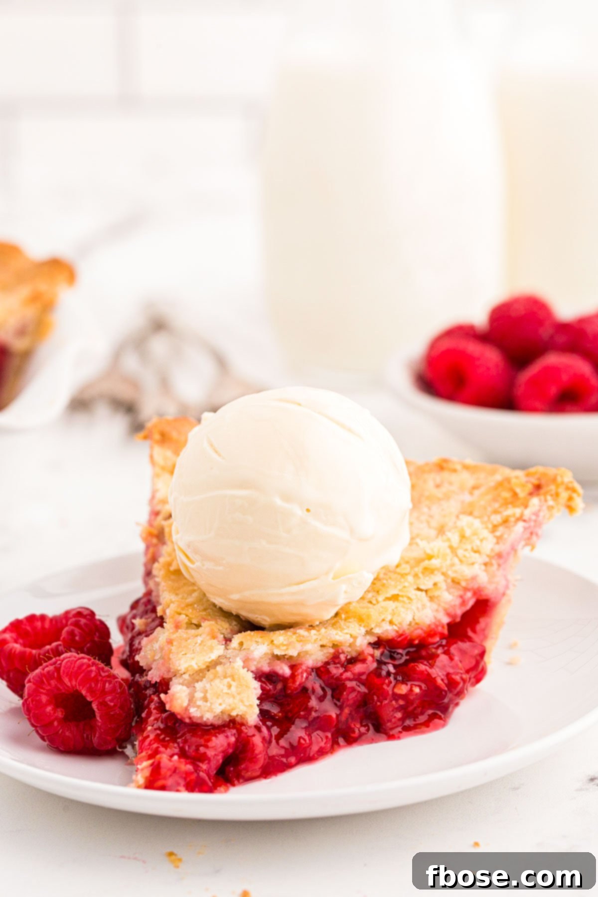A perfectly baked slice of raspberry pie on a white plate, topped with a scoop of melting vanilla ice cream.