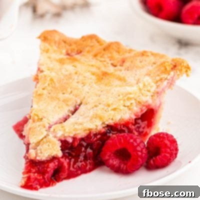 Close up photo of a slice of raspberry pie on a white plate with fresh raspberries around it.