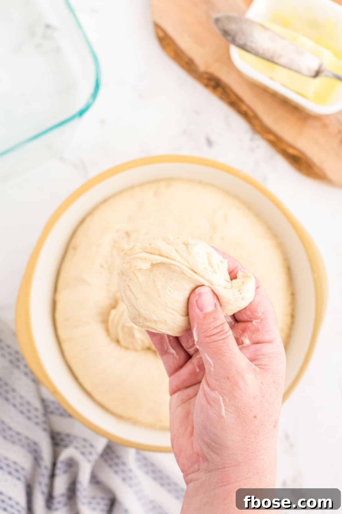 A woman's buttered hand gently pulling a portion of risen sourdough dough from a bowl to begin shaping dinner rolls.