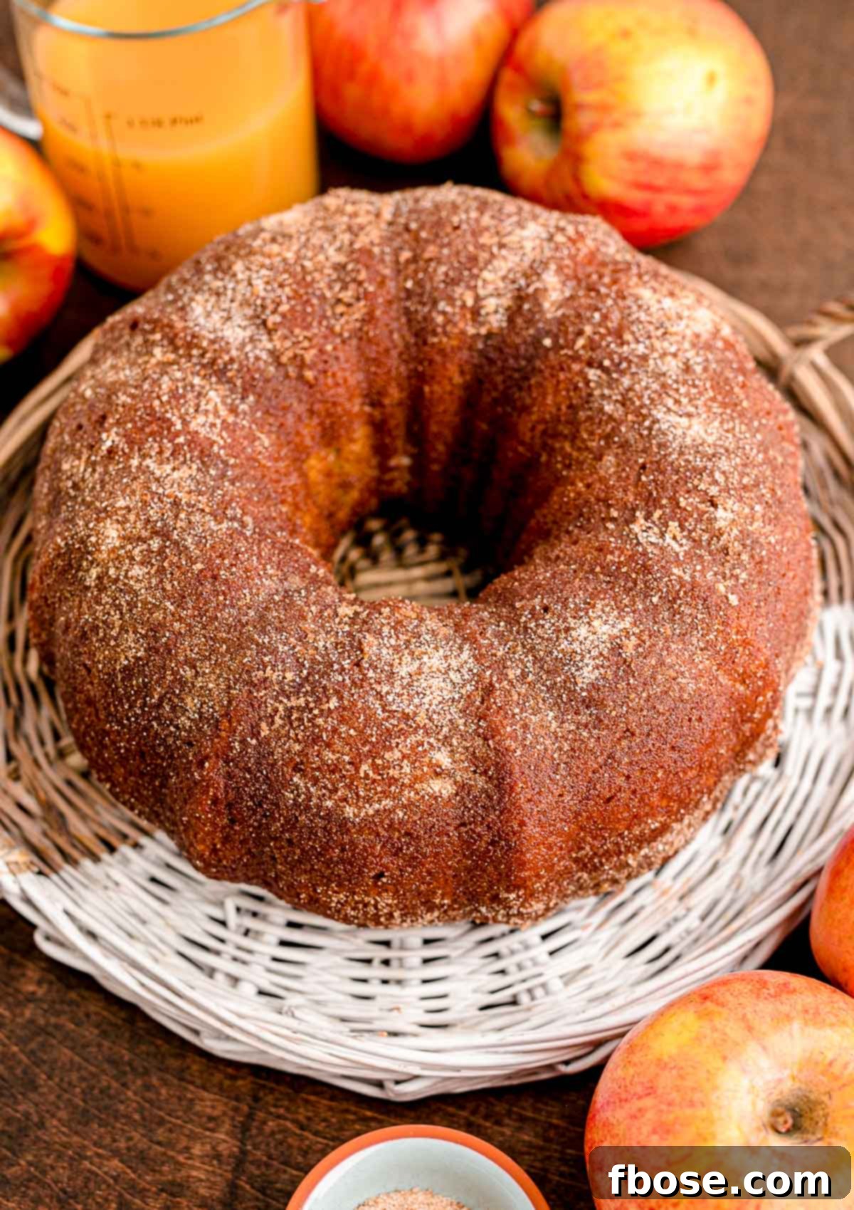 A whole apple cider donut bundt cake on a wicker tray with apples and apple cider in the background.