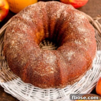 A whole apple cider donut bundt cake on a wicker tray with apples and apple cider in the background.