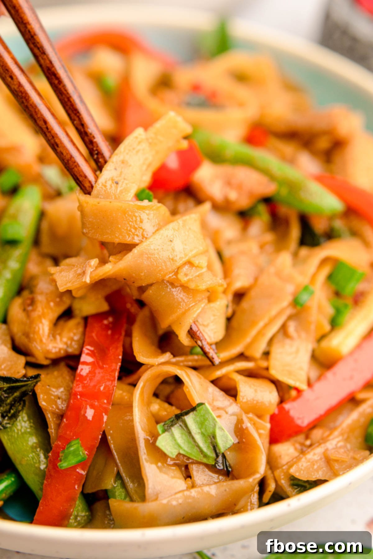 Chopsticks twirling Drunken Noodles from a blue bowl, showcasing the rich sauce and fresh ingredients.