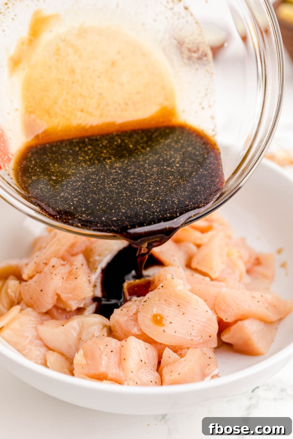 Sauce being poured over pieces of chopped chicken in a white bowl, ready for marinating.