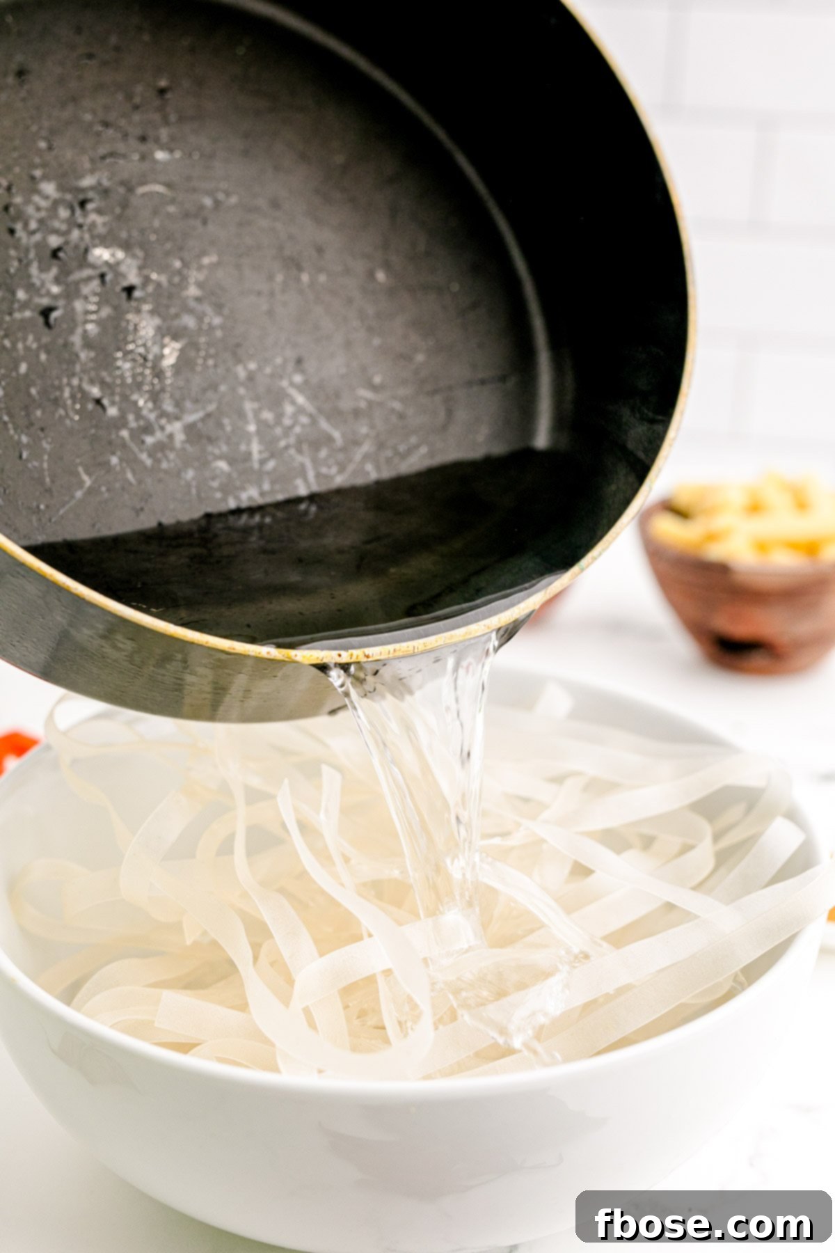Boiling water being poured over flat wide rice noodles in a white bowl, preparing them for the stir-fry.
