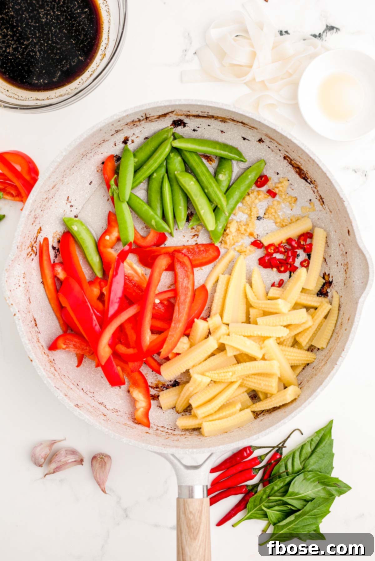 Veggies including garlic, chili, baby corn, red bell pepper, and snow peas added to a hot skillet for stir-frying.