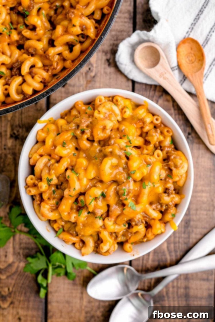 Close up photo of a white bowl with homemade hamburger helper on a wooden table, garnished with fresh parsley, with spoons and linens around it.