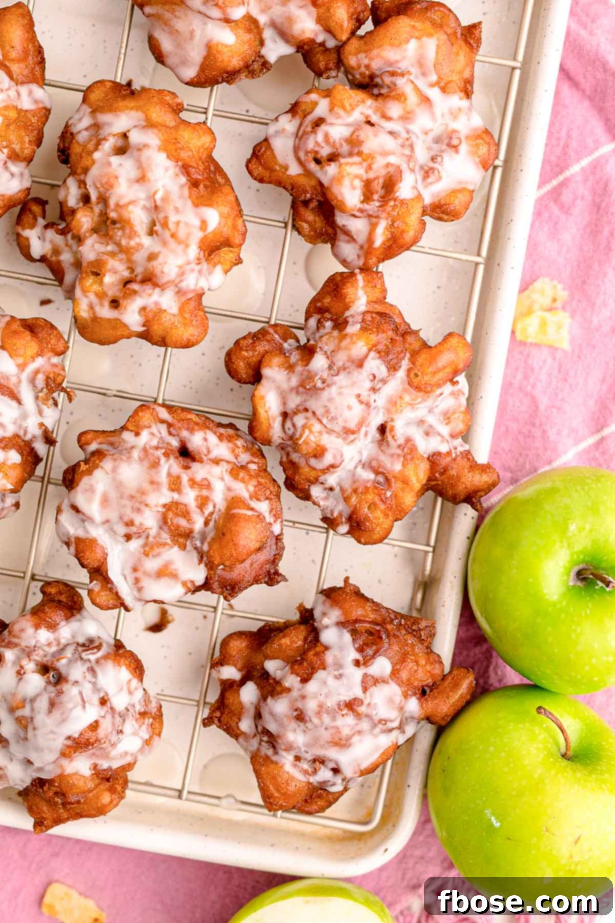 Overhead photo of apple fritters on a baking sheet on a pink napkin with apples on the side.