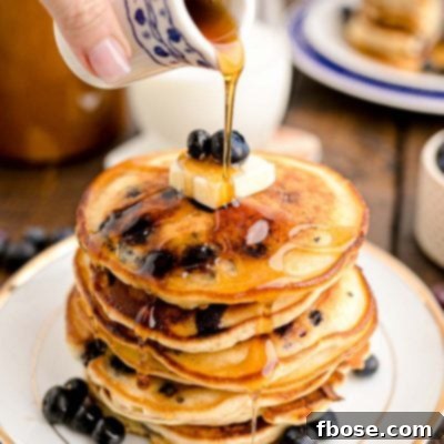 Close up photo of a woman's hand pouring maple syrup over a stack of blueberry pancakes on a white plate on a wooden table.